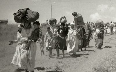 Line of women and children walking along a dusty road, carrying large bundles on their heads amid a refugee-like crowd.