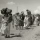 Line of women and children walking along a dusty road, carrying large bundles on their heads amid a refugee-like crowd.