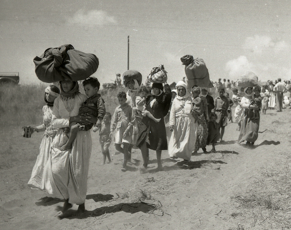 Line of women and children walking along a dusty road, carrying large bundles on their heads amid a refugee-like crowd.
