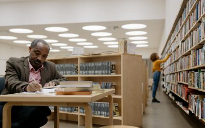 man in brown suit jacket writing on table inside the library