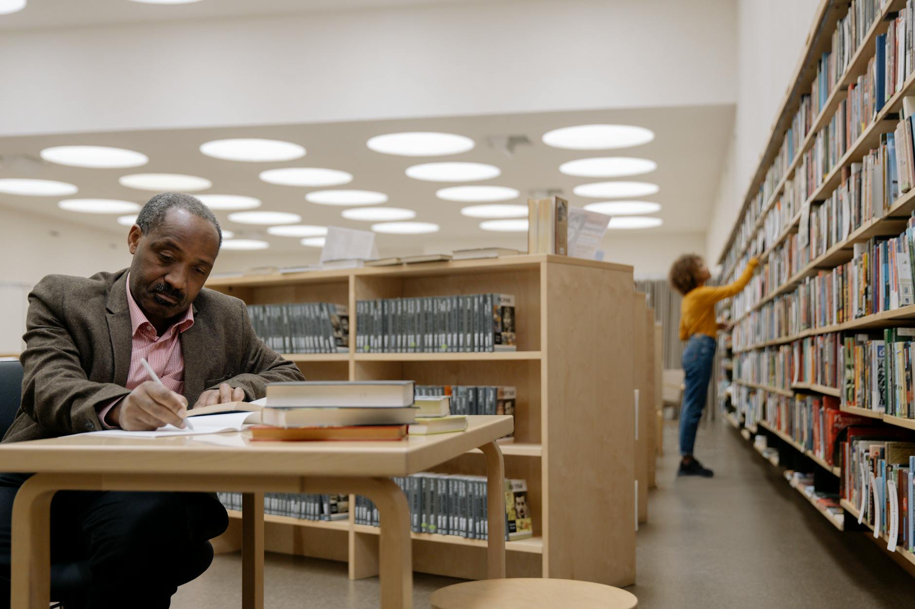 man in brown suit jacket writing on table inside the library